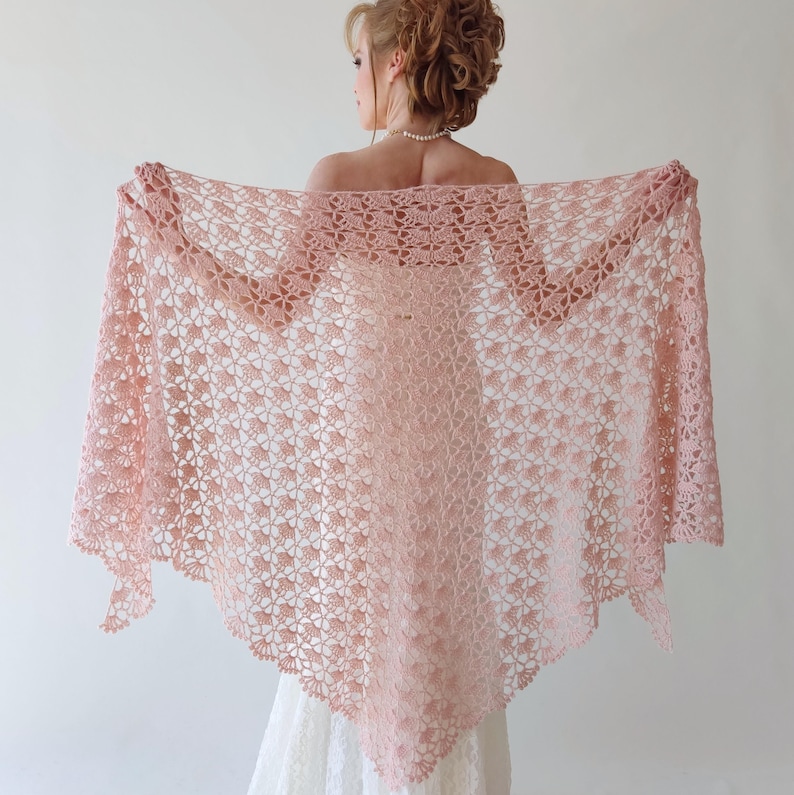 Puede incluir: Un chal rosa claro, de tejido abierto, con un estampado floral. El chal se sostiene abierto, mostrando su delicado dise&ntilde;o y textura. La prenda parece hecha a mano, adecuada para una ocasi&oacute;n especial.