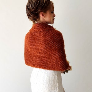 May include: A burnt orange textured knit bolero shrug with a folded collar, worn over a white lace dress. The garment features a dense, fuzzy yarn construction that covers the shoulders and upper back, providing a warm, layered appearance.
