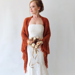 May include: A rust-colored knitted shawl with long fringe edges draped over a white strapless lace dress. The shawl features a textured knit pattern and hangs loosely over the shoulders, paired with a bouquet of white and peach-toned roses.