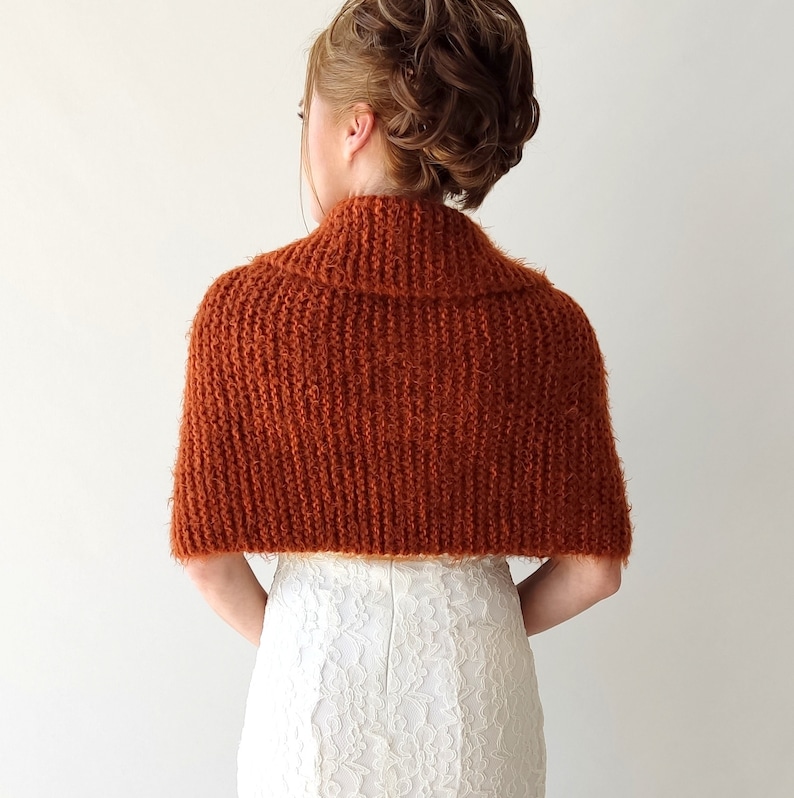 May include: A rust-colored, knitted shrug with a textured, ribbed pattern and a wide collar. The garment covers the shoulders and upper back, featuring a fuzzy, soft-touch yarn construction that provides a layered look over a white dress.