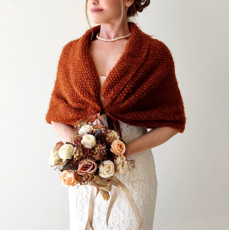 May include: A rust-colored, textured knit shawl with a wide collar and a brown ribbon tie closure. The shawl is worn over a white lace dress, paired with a pearl necklace and a bouquet of white, peach, and brown artificial flowers.