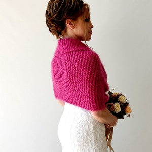 K&ouml;nnte beinhalten: Eine rosafarbene gestrickte Brautjacke mit flauschiger Textur. Die Jacke hat einen Kragen und wird &uuml;ber einem wei&szlig;en Spitzenkleid getragen.