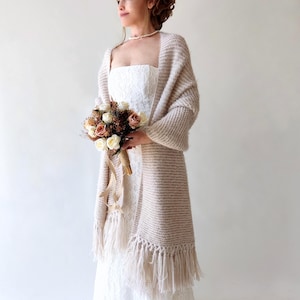 May include: A cream-colored knitted shawl with fringe trim. The shawl is draped over a person wearing a white strapless dress and holding a bouquet of flowers.