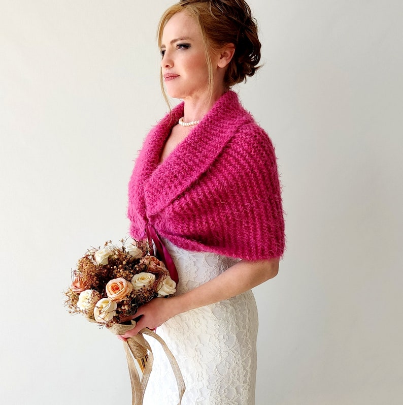 K&ouml;nnte beinhalten: Ein rosafarbener gestrickter Braut-Schal mit einem breiten Kragen und einer Schleife vorne. Der Schal wird &uuml;ber einem wei&szlig;en Spitzenkleid getragen. Die Braut h&auml;lt einen Strau&szlig; cremefarbener und pfirsichfarbener Rosen.