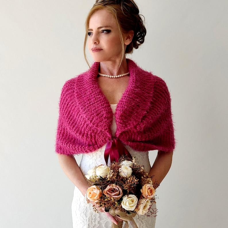 K&ouml;nnte beinhalten: Ein rosa gestrickter Schal mit einer Schleife aus Satinband. Der Schal ist &uuml;ber ein wei&szlig;es Kleid gelegt und das Model h&auml;lt einen Blumenstrau&szlig;.