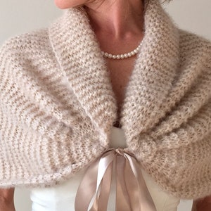 May include: A beige knitted shawl with a satin ribbon tie. The shawl has a soft, fuzzy texture and a wide, open collar.
