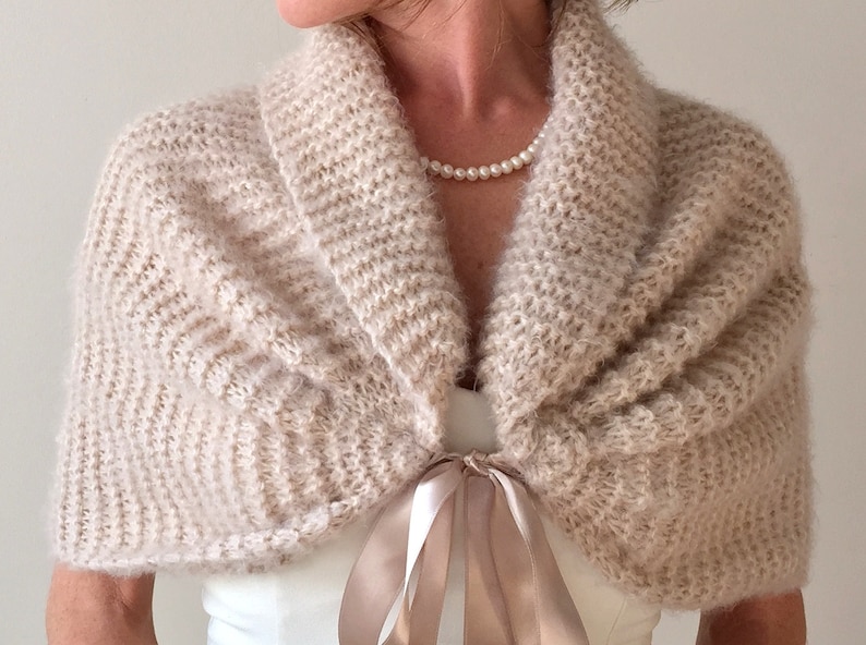 May include: A beige knitted shawl with a satin ribbon tie. The shawl has a soft, fuzzy texture and a wide, open collar.