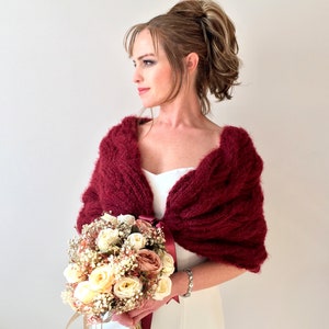 May include: A burgundy knitted shawl with a cable knit pattern. The shawl is draped over the shoulders of a person holding a bouquet of white and cream flowers.