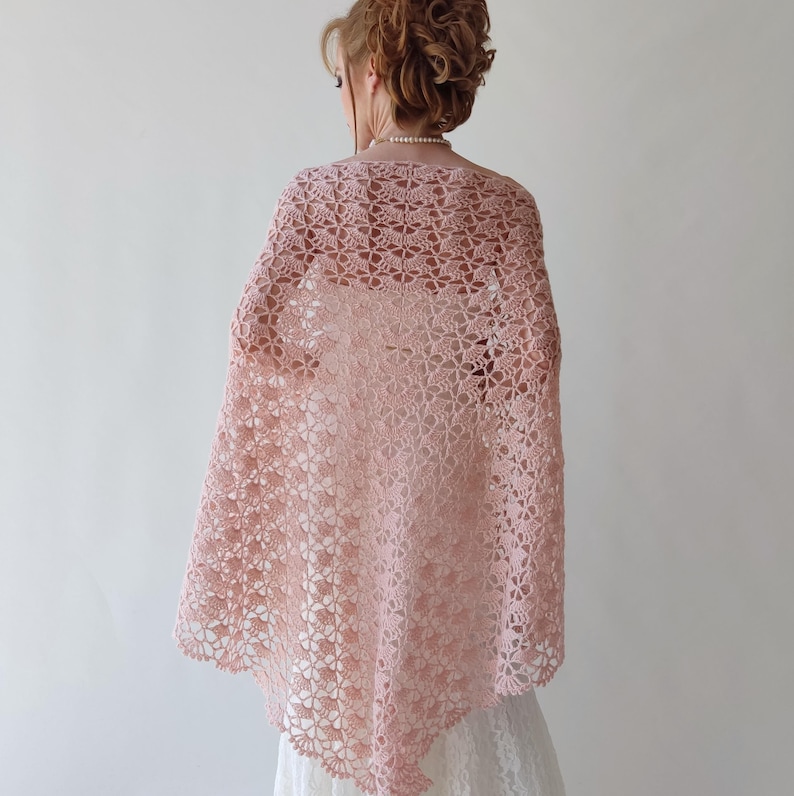 Puede incluir: Un chal rosa claro, de tejido abierto, con un dise&ntilde;o floral. El chal est&aacute; drapeado sobre los hombros, creando un aspecto delicado y aireado. El intrincado dise&ntilde;o de ganchillo a&ntilde;ade un toque de elegancia a la prenda. El chal probablemente est&aacute; hecho de un material suave y ligero.