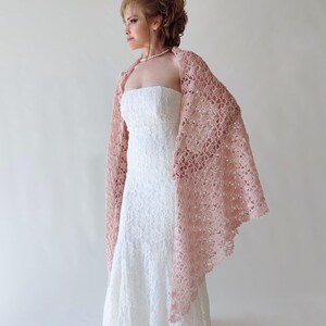 Puede incluir: Un chal de ganchillo rosa claro con un patr&oacute;n floral calado. Se lleva sobre un vestido blanco de encaje sin tirantes. El dise&ntilde;o delicado y el color suave del chal crean un look elegante y femenino, adecuado para bodas u ocasiones especiales.