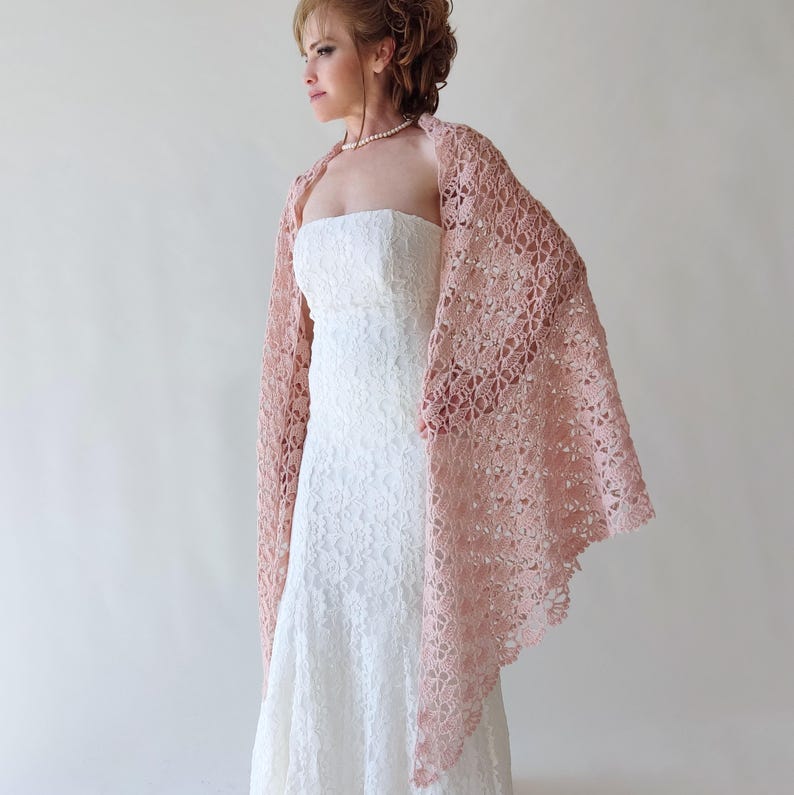 Puede incluir: Un chal de ganchillo rosa claro con un patr&oacute;n floral calado. Se lleva sobre un vestido blanco de encaje sin tirantes. El dise&ntilde;o delicado y el color suave del chal crean un look elegante y femenino, adecuado para bodas u ocasiones especiales.