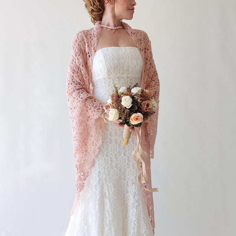 Puede incluir: Un chal de encaje rosa claro sobre un vestido de novia blanco. El chal tiene un delicado dise&ntilde;o floral. La novia sostiene un ramo de flores blancas, melocot&oacute;n y marr&oacute;n, atado con hilo y cintas. El fondo es blanco liso.