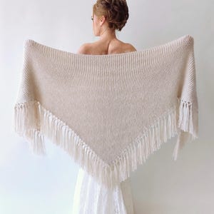 May include: A triangular cream-colored knit shawl featuring a textured stitch pattern and long, dangling fringe along the bottom edges. The garment is draped horizontally, showcasing the soft, woven fabric and the uniform, knotted tassel details.