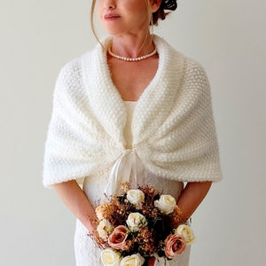 May include: White knitted bridal shawl with a satin ribbon tie. The shawl is draped over the shoulders and tied at the front. The shawl is made of a soft, fluffy yarn.
