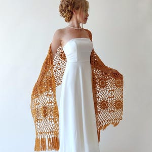 May include: A mustard yellow crochet shawl with a delicate floral pattern and fringe trim. The shawl is draped over a white strapless dress.