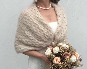 May include: A beige knitted shawl with a delicate openwork pattern. The shawl is draped over the shoulders of a person wearing a white dress and holding a bouquet of dried flowers.
