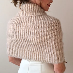 May include: A soft, light beige knitted shrug with a turtleneck collar. The shrug is made of a fuzzy yarn and has a loose, flowing fit.