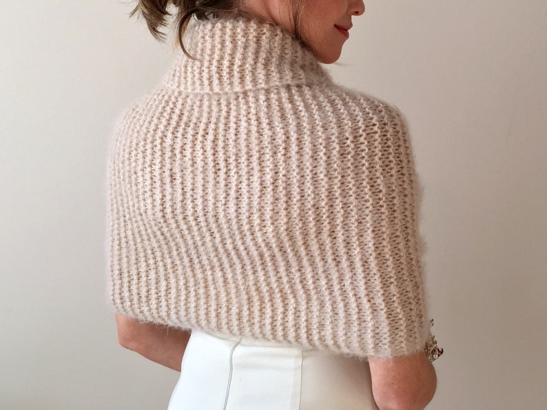 May include: A soft, light beige knitted shrug with a turtleneck collar. The shrug is made of a fuzzy yarn and has a loose, flowing fit.