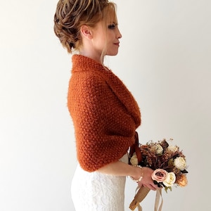 May include: A textured, burnt orange knit bolero shrug with a wide collar, worn over a white lace dress. The garment features a fuzzy, nubby yarn construction and is paired with a bouquet of cream, peach, and brown artificial flowers.