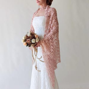 Puede incluir: Un chal de ganchillo rosa claro de tejido abierto con mangas largas. El chal est&aacute; drapeado sobre un vestido blanco sin tirantes. Un ramo de flores con rosas melocot&oacute;n y blancas se sostiene en las manos.