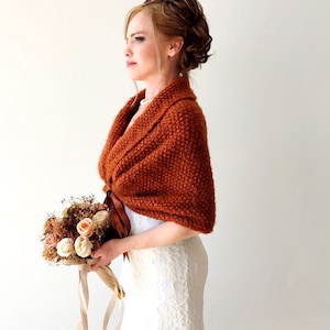 May include: A rust-colored knitted shawl with a textured surface and a brown ribbon tie closure. The shawl is worn over a white lace garment and is paired with a bouquet of dried flowers featuring cream and peach-colored roses.