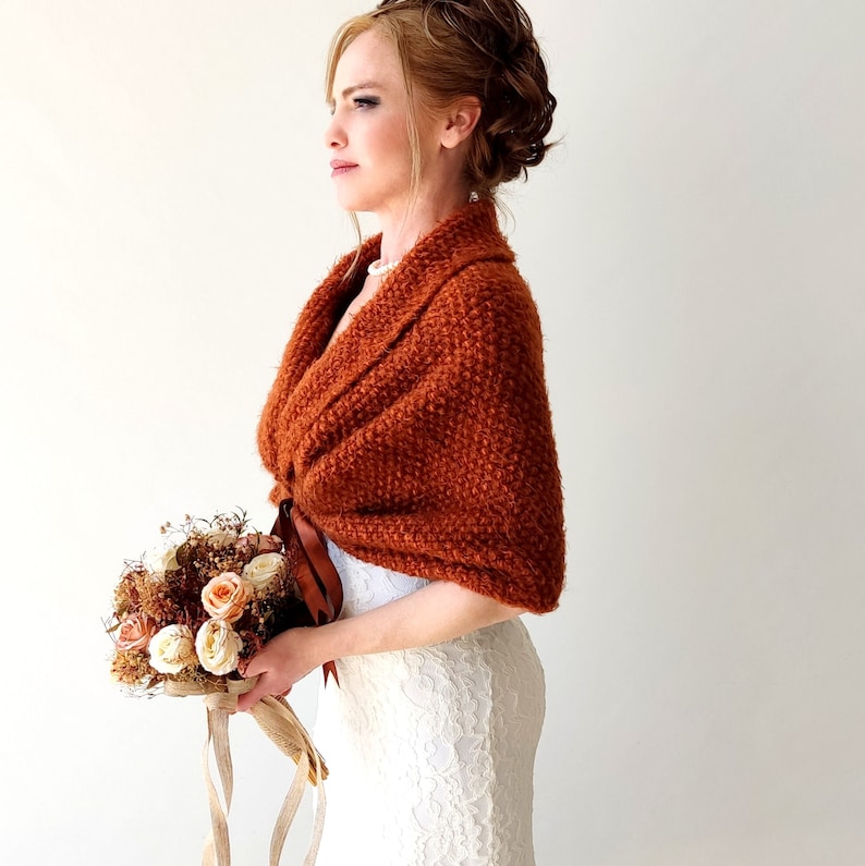 May include: A rust-colored knitted shawl with a textured surface and a brown ribbon tie closure. The shawl is worn over a white lace garment and is paired with a bouquet of dried flowers featuring cream and peach-colored roses.