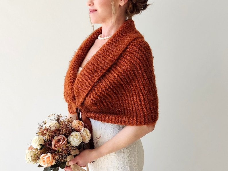 May include: A rust-colored knitted shrug featuring a wide collar and textured yarn construction. The garment is worn over a white lace dress and is paired with a bouquet of artificial roses in shades of cream, peach, and brown.