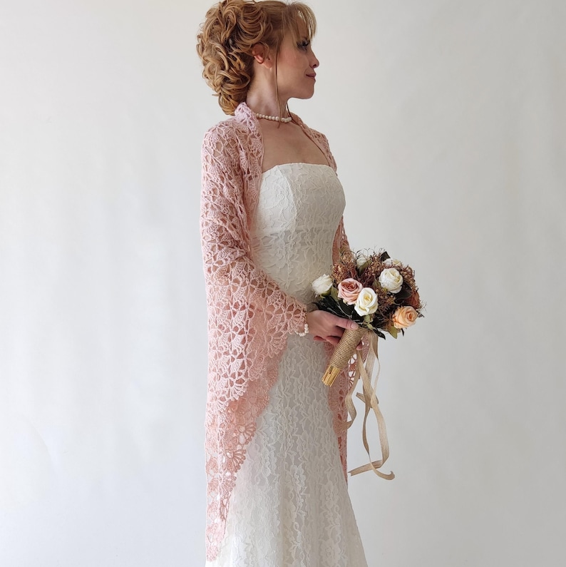 Puede incluir: Una novia con un vestido de novia de encaje blanco con un chal de encaje rosa. Sostiene un ramo de flores con rosas beige y rosas. El chal tiene mangas largas y un delicado estampado floral.