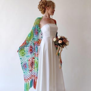 Peut inclure: Une femme portant une robe blanche sans bretelles avec un châle en crochet coloré. Le châle a une frange et présente un motif floral dans les tons de vert, bleu, orange et rouge. Elle tient un bouquet de fleurs.