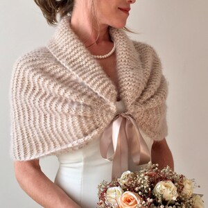 May include: A blush-colored knitted bridal shawl with a satin ribbon tie. The shawl is draped over a white wedding dress and the bride is holding a bouquet of dried flowers.