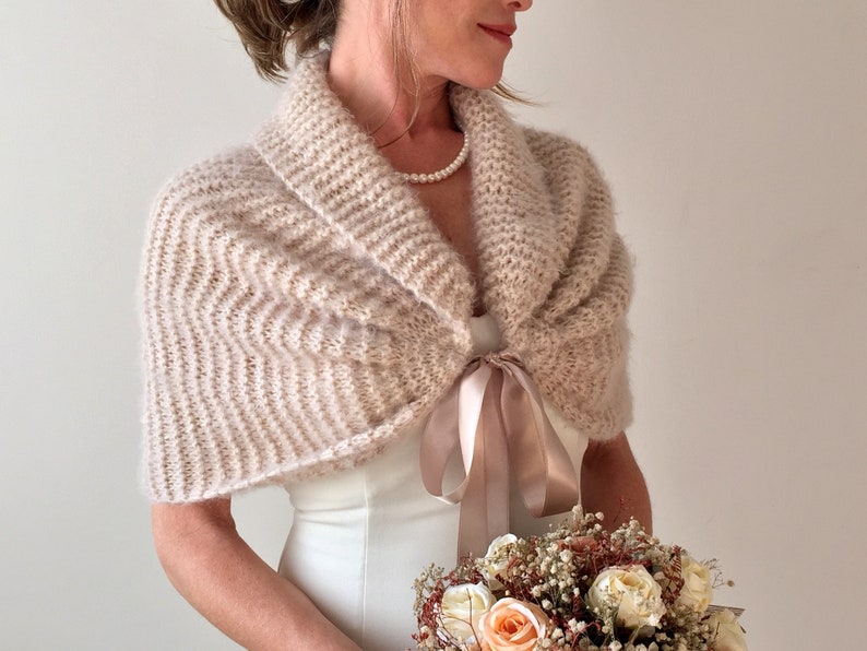 May include: A blush-colored knitted bridal shawl with a satin ribbon tie. The shawl is draped over a white wedding dress and the bride is holding a bouquet of dried flowers.
