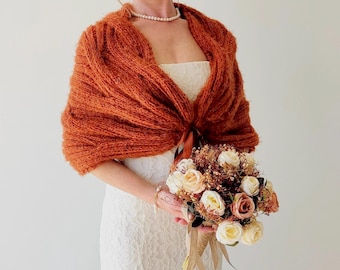 Puede incluir: Un chal de punto naranja quemado con un lazo de cinta de satén. El chal está drapeado sobre un vestido de encaje blanco y la modelo sostiene un ramo de flores secas.