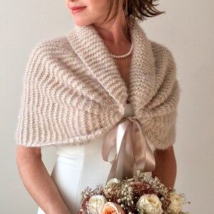 May include: A blush-colored knitted shawl with a satin ribbon tie. The shawl is draped over a white dress and the model is holding a bouquet of flowers.