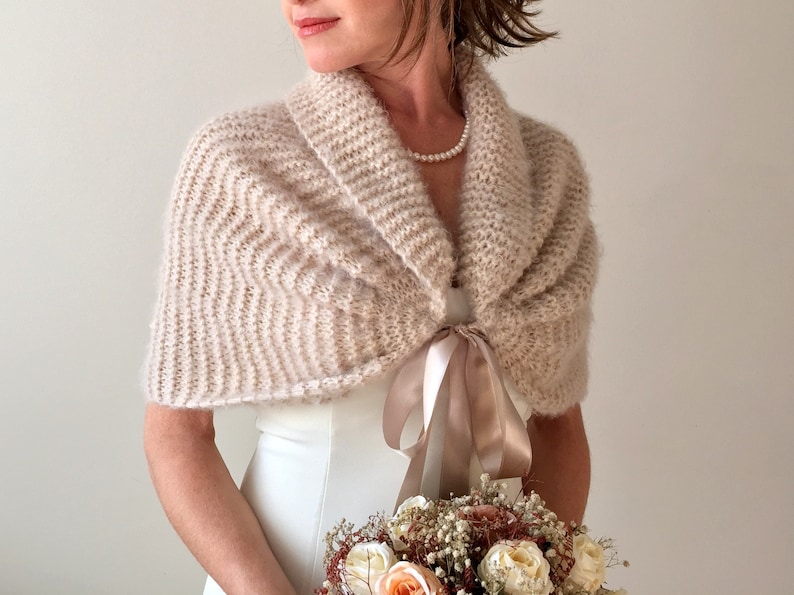 May include: A blush-colored knitted shawl with a satin ribbon tie. The shawl is draped over a white dress and the model is holding a bouquet of flowers.