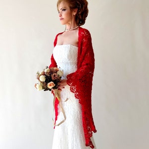 May include: A bride in a white lace wedding dress with a red, open-weave crochet shawl. She holds a bouquet of flowers with a ribbon. The shawl has a scalloped edge and covers her shoulders and arms.