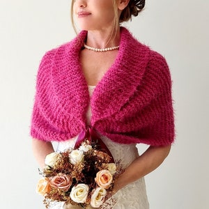 K&ouml;nnte beinhalten: Ein rosa gestrickter Schal mit einer burgunderfarbenen Schleife. Der Schal wird &uuml;ber einem wei&szlig;en Kleid getragen und die Person h&auml;lt einen Blumenstrau&szlig;.