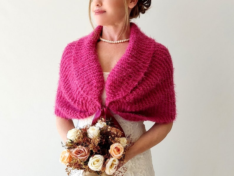K&ouml;nnte beinhalten: Ein rosa gestrickter Schal mit einer burgunderfarbenen Schleife. Der Schal wird &uuml;ber einem wei&szlig;en Kleid getragen und die Person h&auml;lt einen Blumenstrau&szlig;.