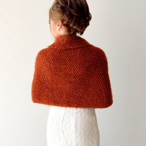 May include: A rust-colored, textured knit shawl with a folded collar draped over the shoulders. The garment features a dense, fuzzy yarn construction with a consistent woven pattern, providing full coverage across the upper back and shoulders.