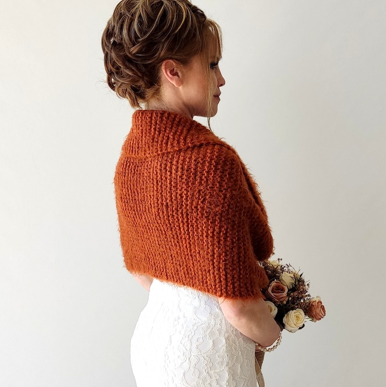May include: A rust-colored, knitted shrug with a textured, fuzzy yarn finish and a wide collar. The garment features a ribbed knit pattern and covers the shoulders and upper back, worn over a white lace dress with a floral bouquet in hand.
