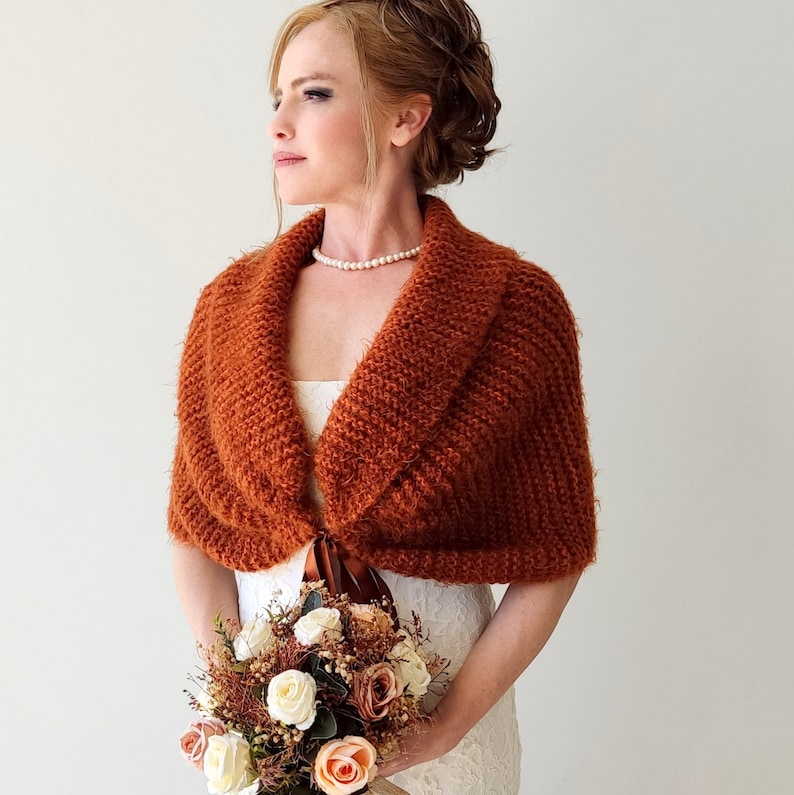 May include: A rust-colored knitted shawl with a wide collar and a textured surface, featuring a brown ribbon tie closure at the front. The garment is worn over a white dress and is paired with a bouquet of white, peach, and brown artificial roses.