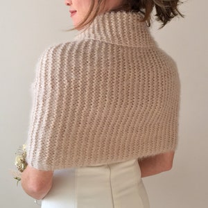 May include: A soft, blush-colored knitted shrug with a turtleneck collar. The shrug is made of a fine yarn and has a delicate, feminine look.