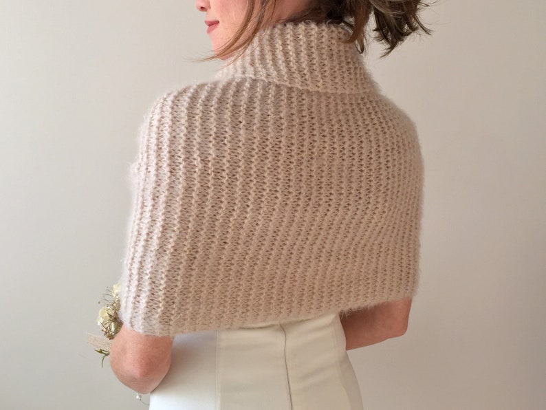 May include: A soft, blush-colored knitted shrug with a turtleneck collar. The shrug is made of a fine yarn and has a delicate, feminine look.