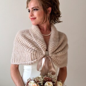 May include: A beige knitted bridal shawl with a satin ribbon tie. The shawl is draped over the shoulders of a person wearing a white dress.