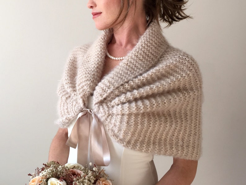 May include: A soft, ivory knitted shawl with a satin ribbon tie. The shawl is draped over the shoulders of a person wearing a white dress.