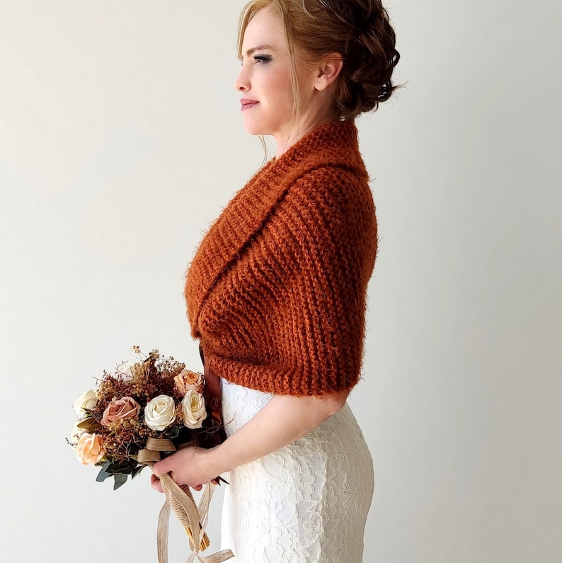 May include: A rust-colored, knitted shrug with a textured, ribbed pattern and a wide collar, worn over a white, lace-textured dress. The garment is paired with a bouquet of cream, peach, and brown dried flowers tied with a tan ribbon.