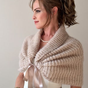 May include: A beige knitted shawl with a satin ribbon tie. The shawl is draped over the shoulders of a person wearing a white dress.