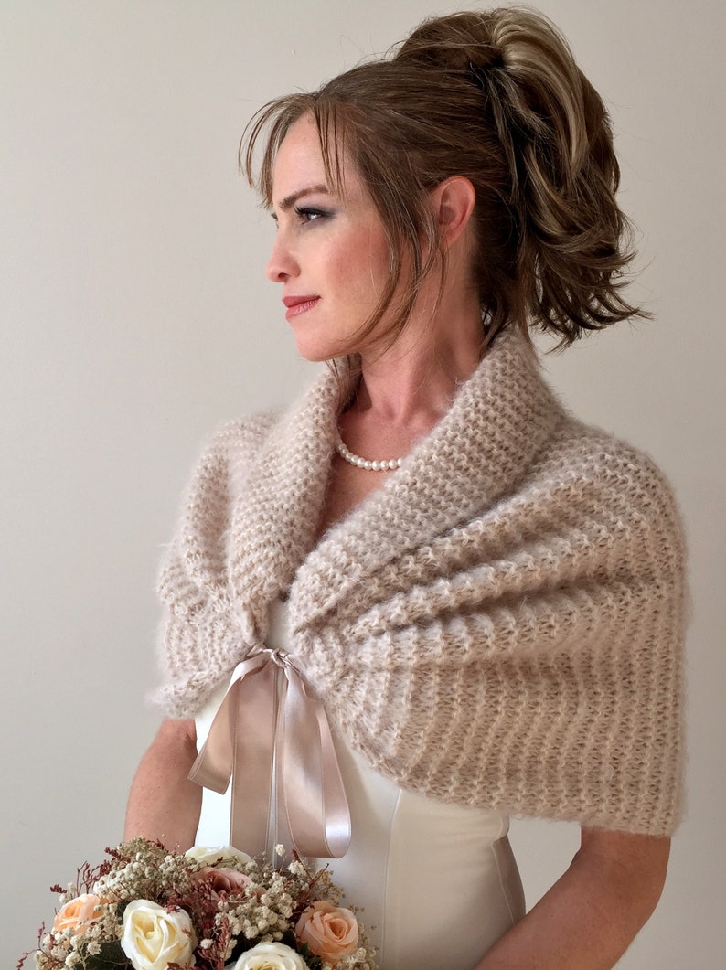 May include: A beige knitted shawl with a satin ribbon tie. The shawl is draped over the shoulders of a person wearing a white dress.
