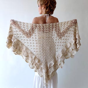 May include: A beige, open-weave crochet shawl with a scalloped edge. The shawl is held open, showcasing its intricate floral pattern and delicate texture. The garment is draped over a person's shoulders.