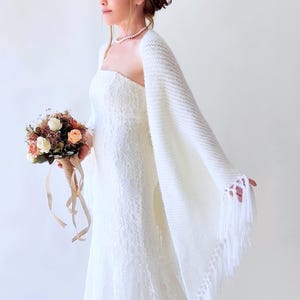May include: A white strapless lace gown paired with a matching white knitted shawl featuring long fringe trim. The ensemble is accessorised with a simple pearl necklace and a bouquet of dried flowers with peach and white roses tied with ribbons.