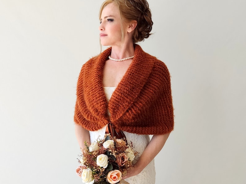May include: A rust-colored knitted shawl with a wide collar and a textured surface, secured at the front with a brown ribbon tie. The garment is worn over a white dress, paired with a pearl necklace and a bouquet of white and peach roses.